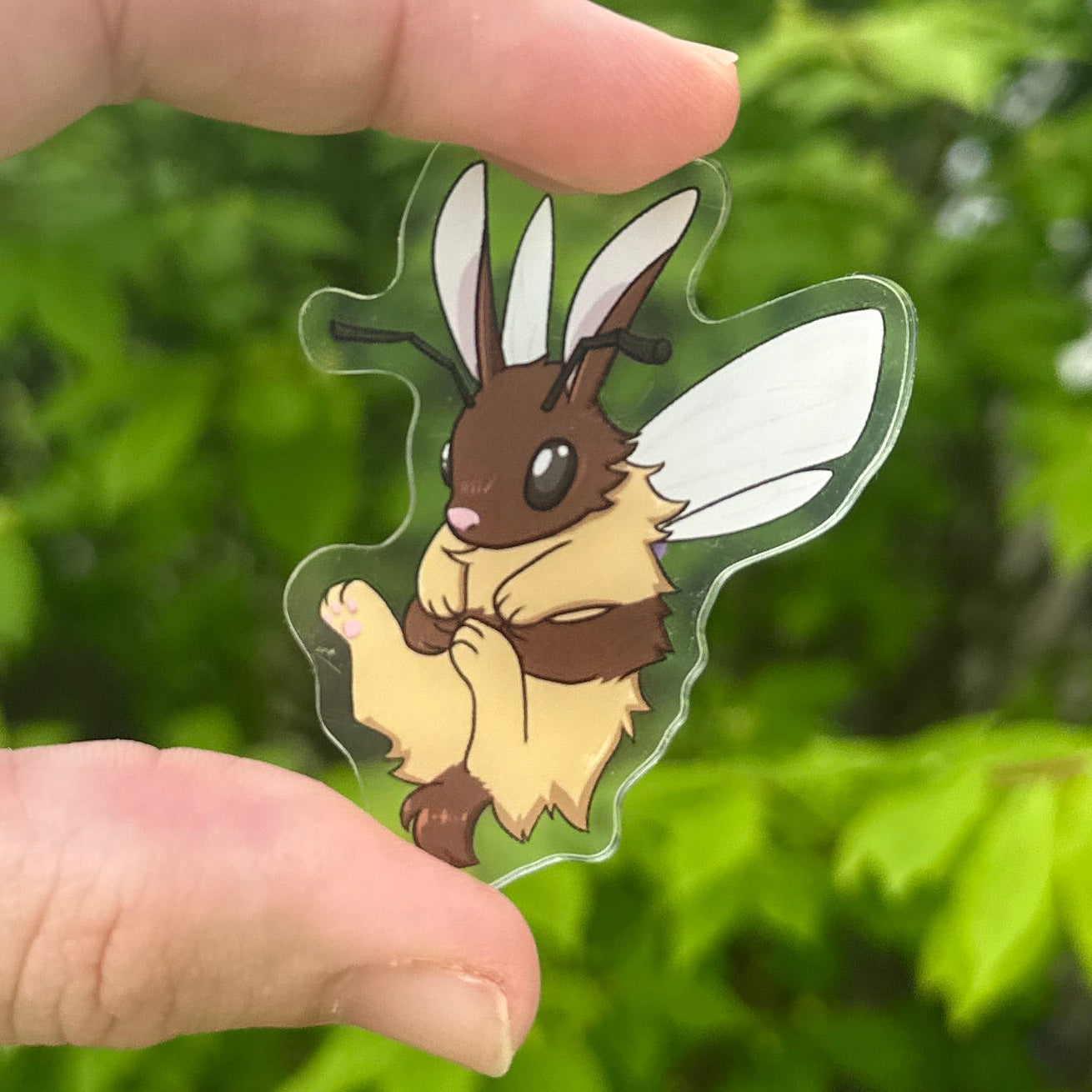 Hand holding a clear pin with bee character against a green leafy background