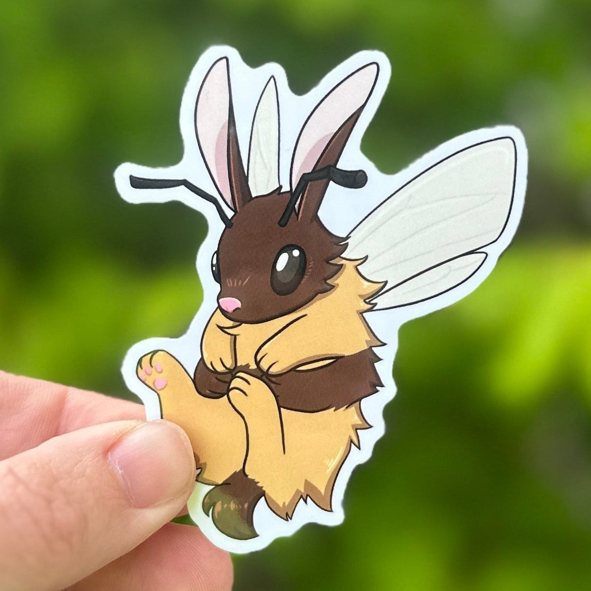 Hand holding a cartoon bee sticker against a green leafy background
