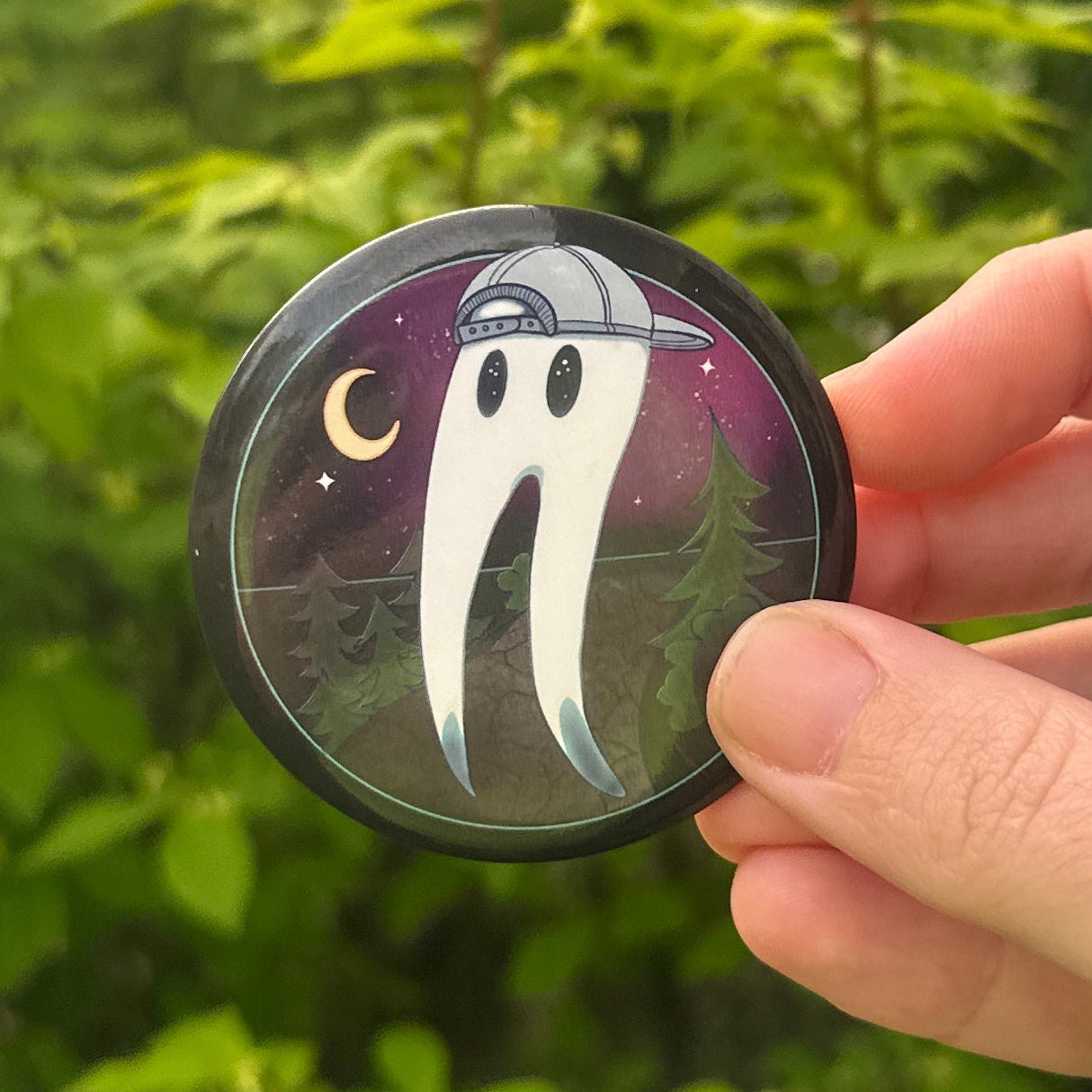 Hand holding a small round pin with a Fresno Nightcrawler design against a green outdoor background