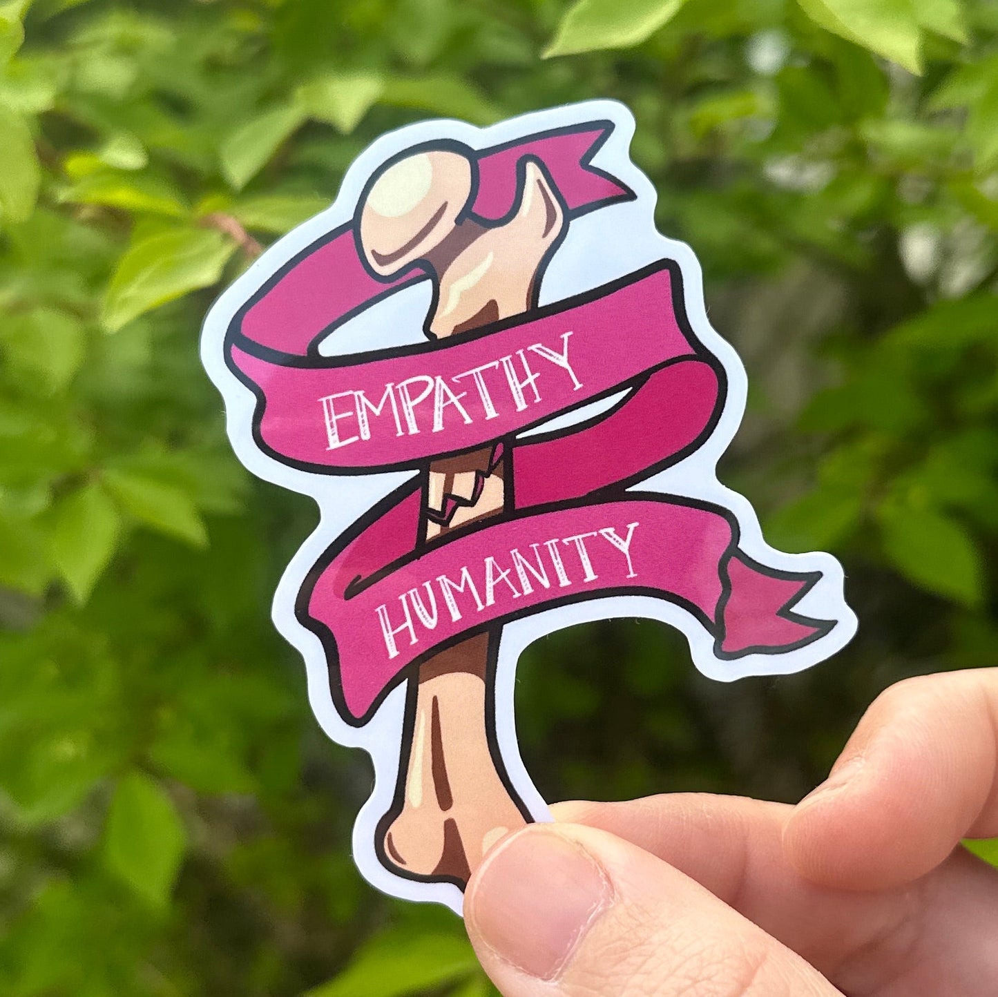Hand holding a pink and white femur and ribbonsticker with text against a green leafy background