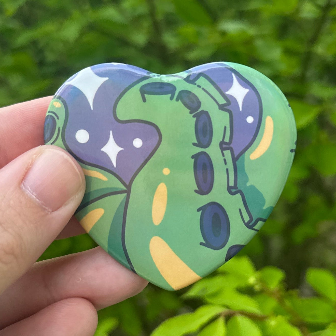 Hand holding a colorful heart-shaped pin with tentacles against a green leafy background