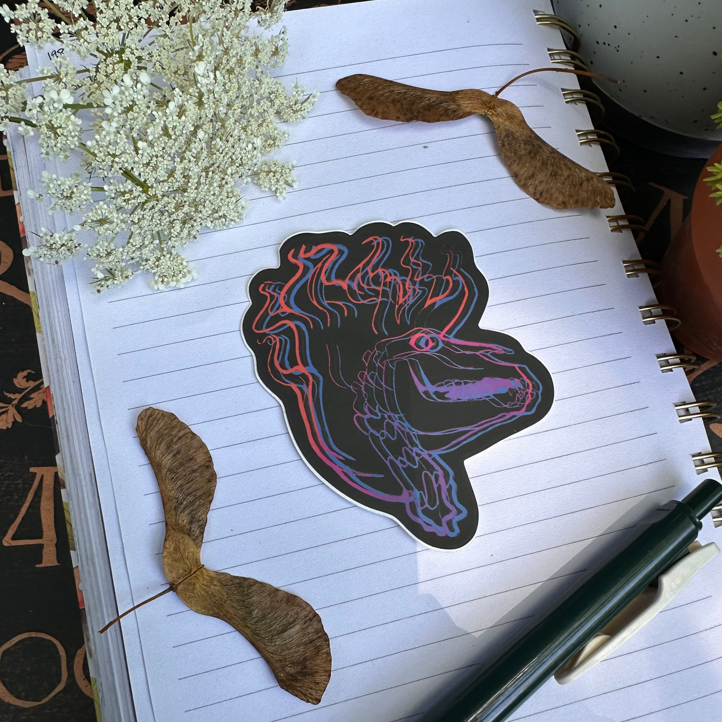 Sticker featuring a horse skull with a flaming mane outlined in purple and pink. Laying on a journal surrounded by flowers.