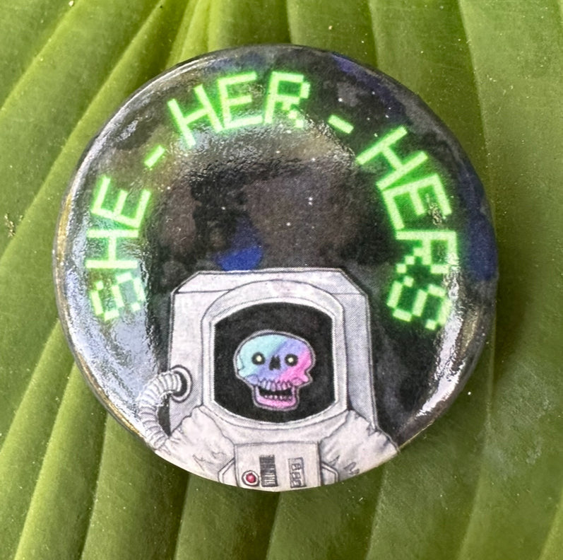 A pin featuring an astronaut motif and the text 'she her hers’ in a stylized font.