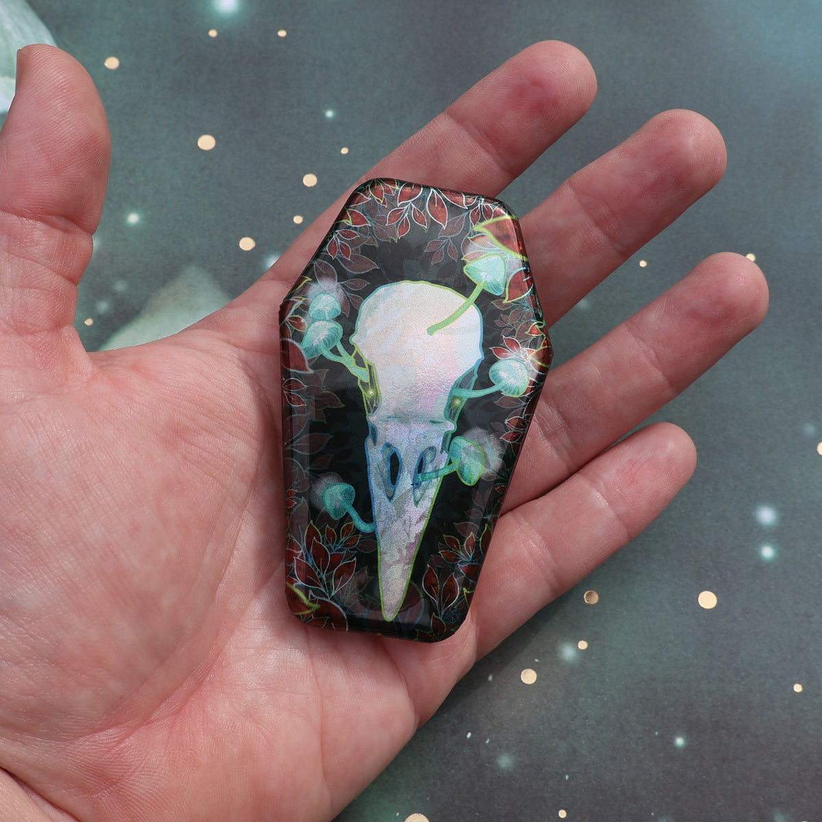 Hand holding the raven skull pin with mushrooms pin for scale
