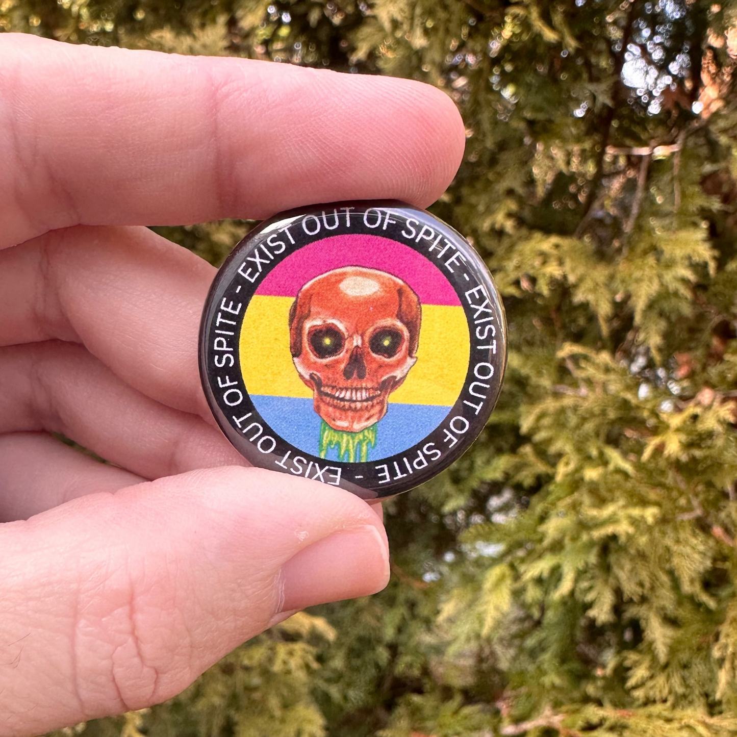 Round pin with a skull over a pansexual pride flag encircled by the text exist out of spite