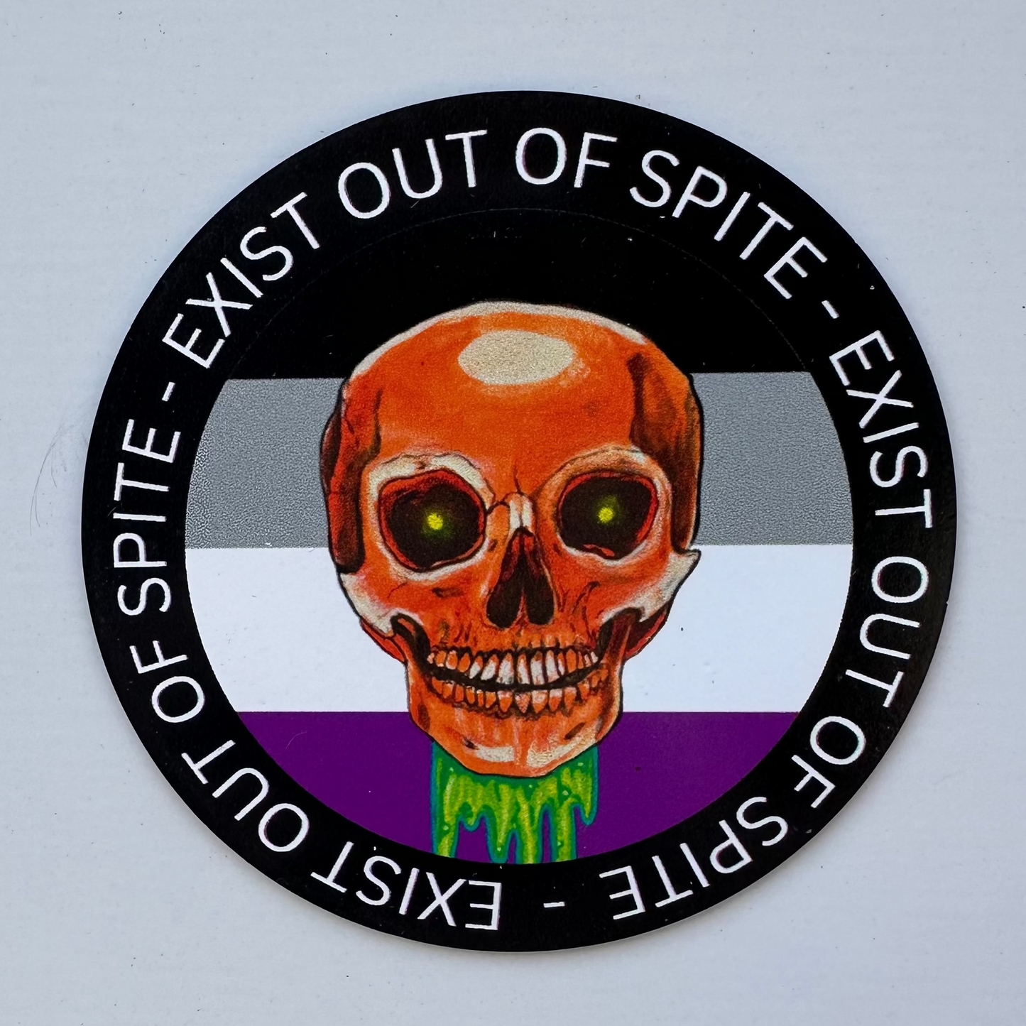 Round magnet with a skull over a asexual pride flag encircled by the text exist out of spite