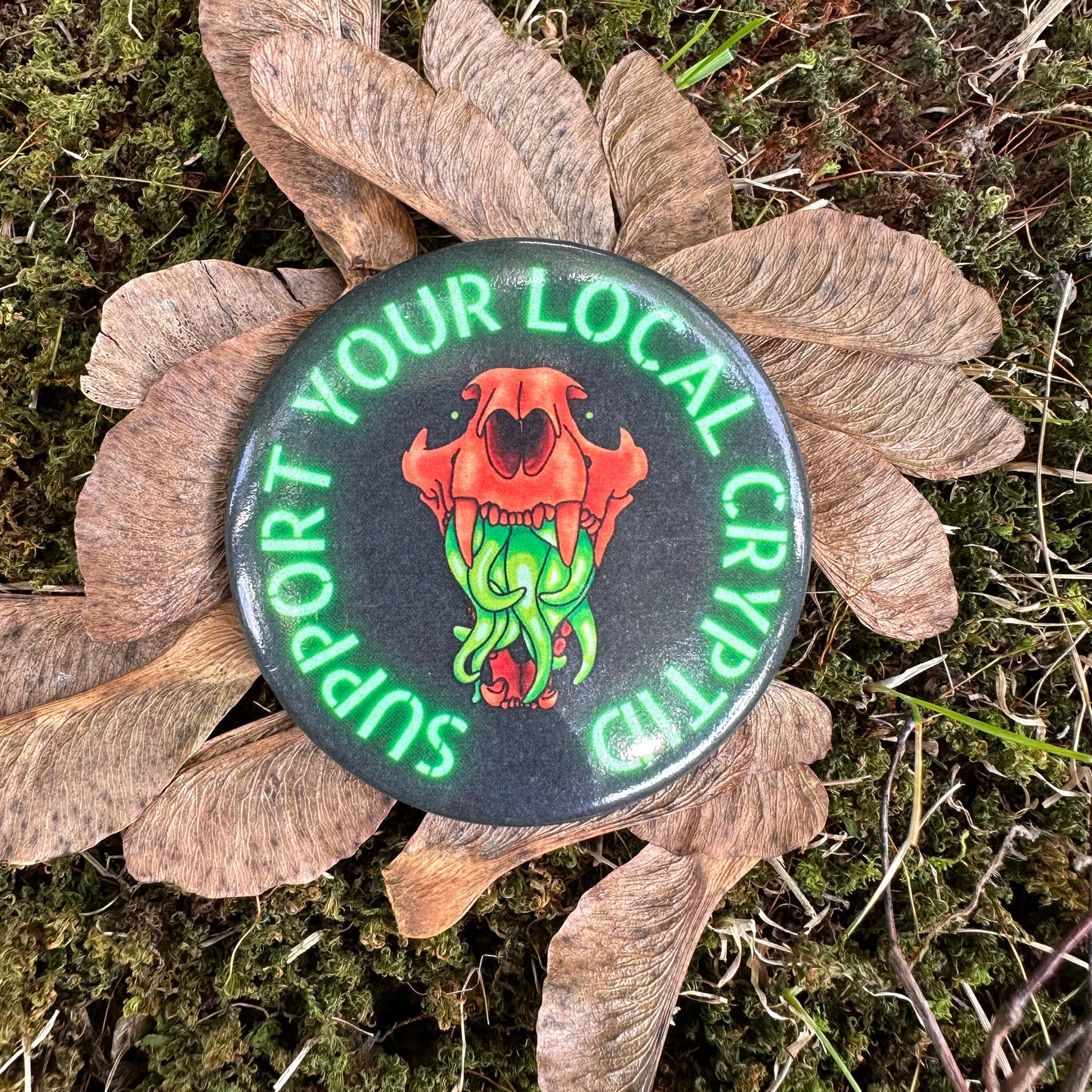 A pin featuring a stylized illustration of an animal skull with vibrant green tentacles, and the text ‘support your local Cryptid' around it.