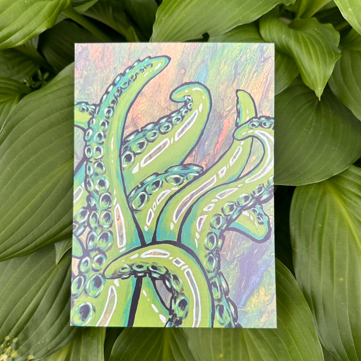 A colorful art print featuring an intricate illustration of tentacles on a glossy cardstock background, presented against a natural green leaf backdrop.