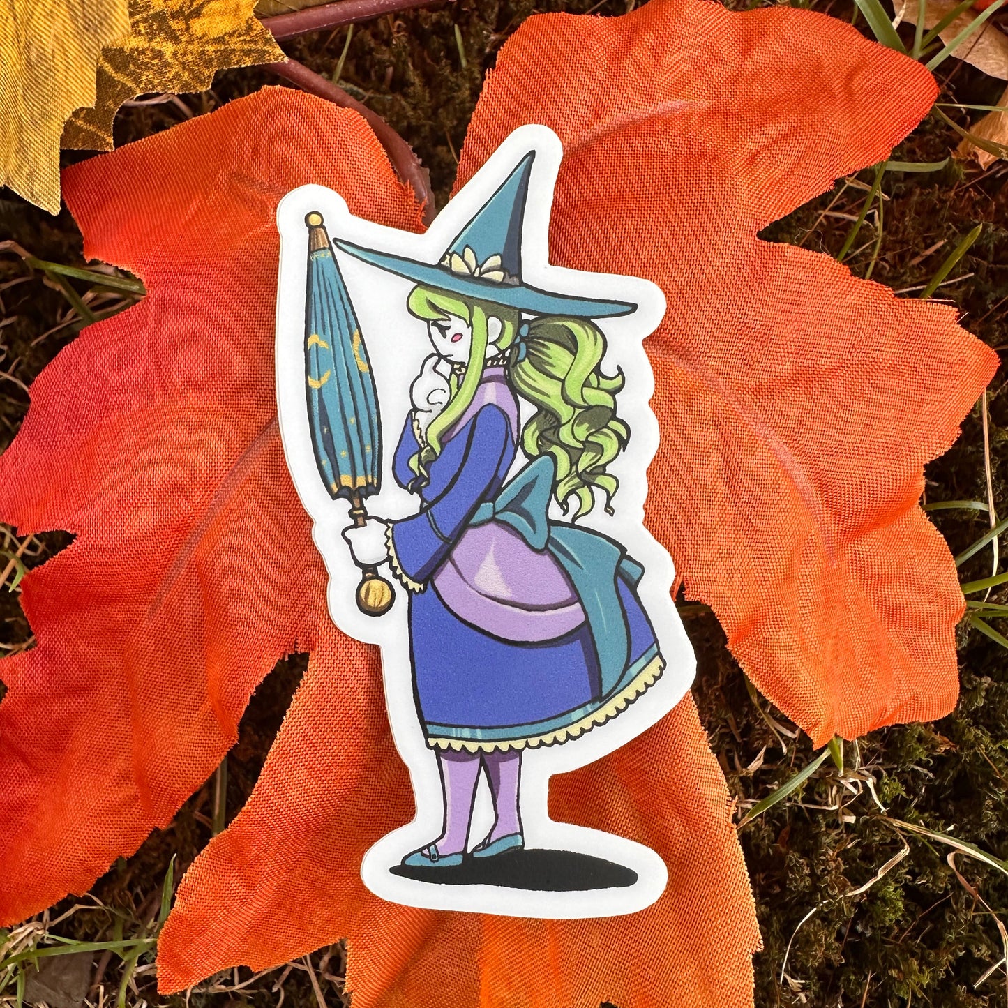 A sticker depicting a sad witch with a green hat and blue dress, standing on a leaf with a autumn background.