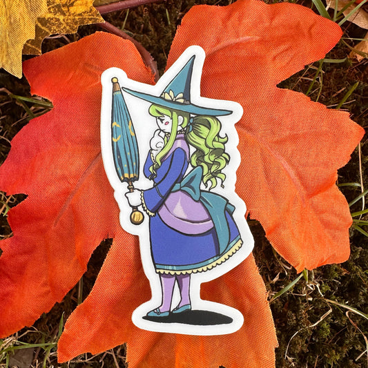 A sticker depicting a sad witch with a green hat and blue dress, standing on a leaf with a autumn background.