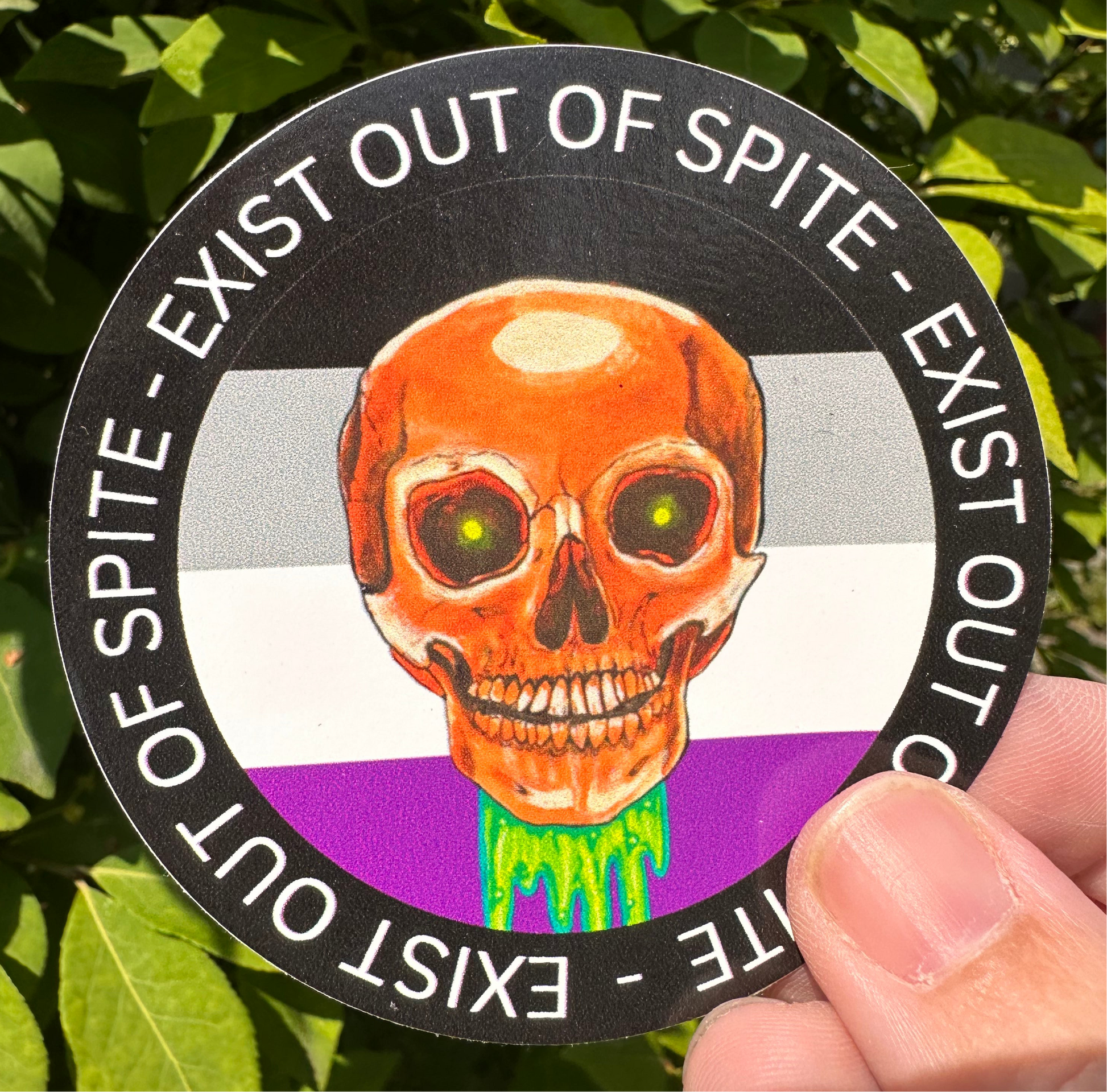 Round sticker with a skull over an asexual pride flag encircled by the text exist out of spite
