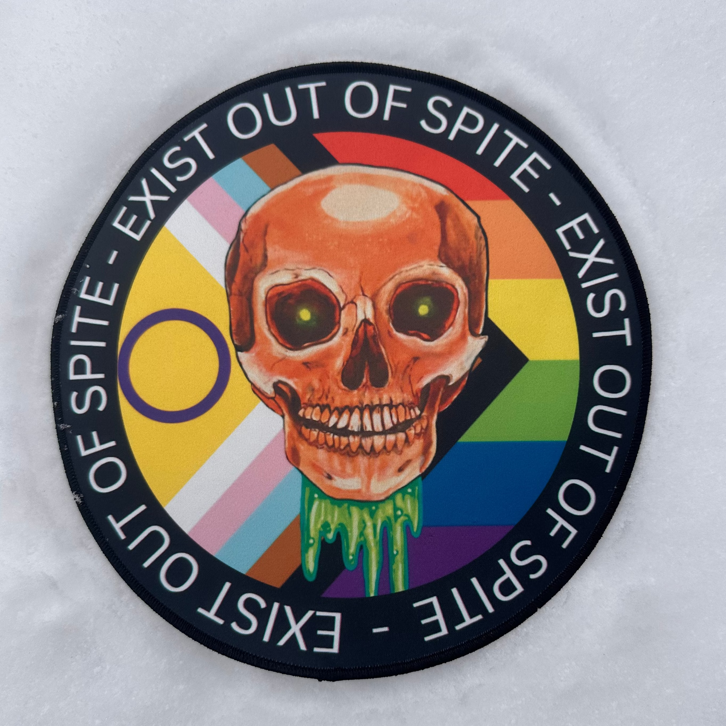 9 inch patch laying in a snow scape featuring a dripping skull in the center, with a rainbow pride flag behind, and the words "Exist Out of Spite" encircling it.