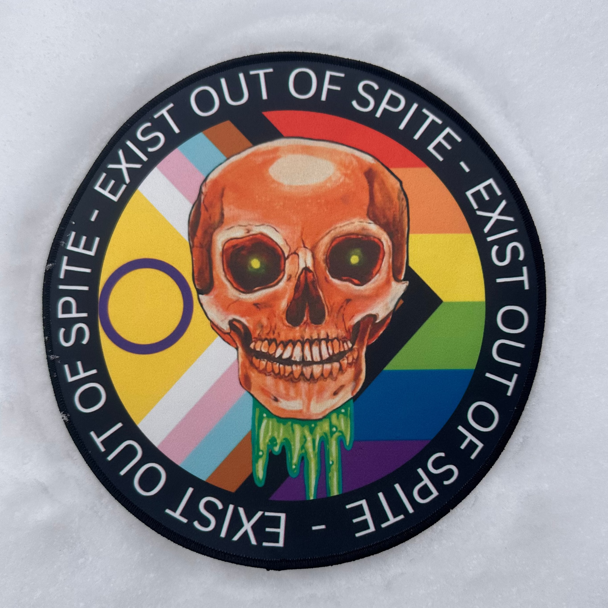 9 inch patch laying in a snow scape featuring a dripping skull in the center, with a rainbow pride flag behind, and the words "Exist Out of Spite" encircling it.