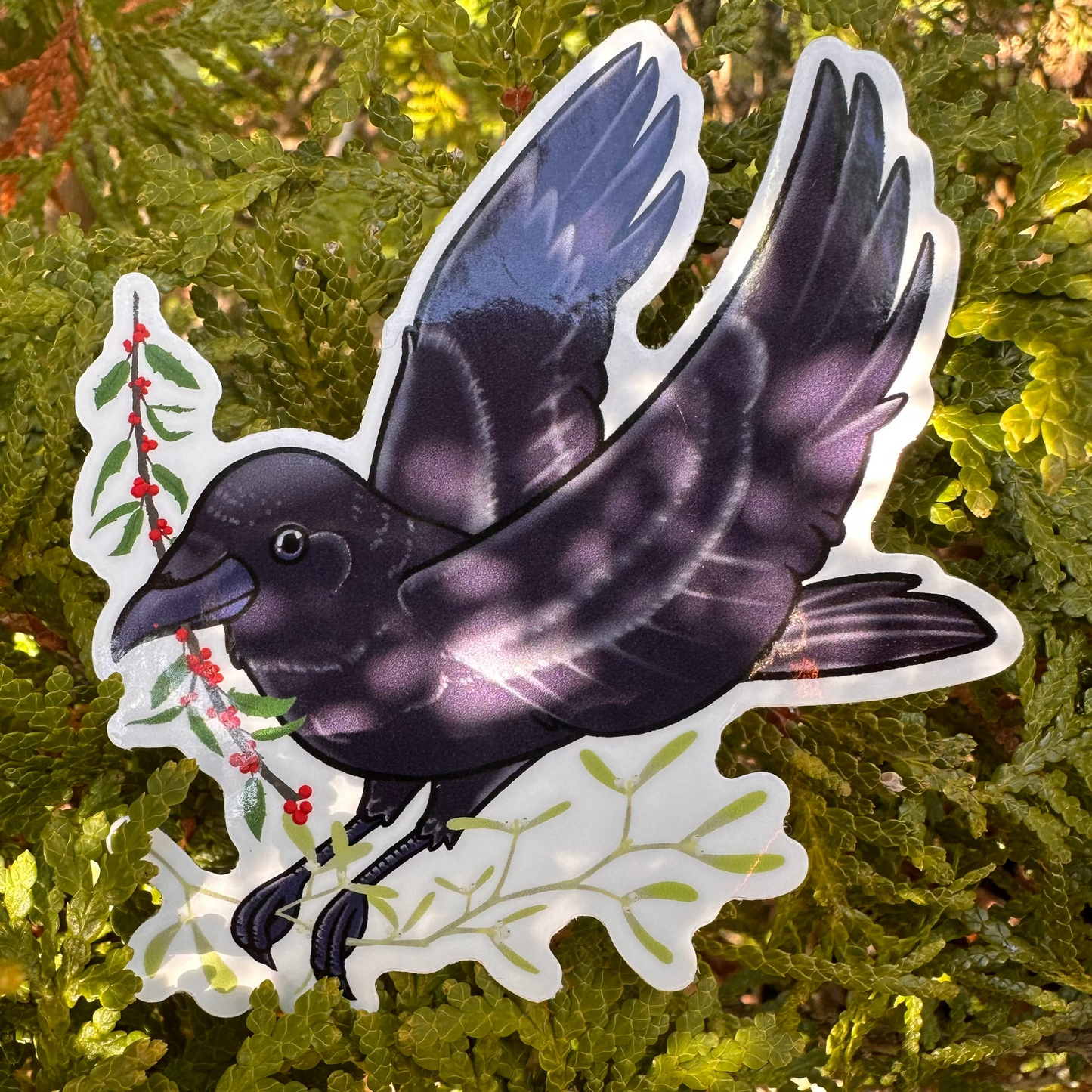 A vinyl sticker depicting a black crow holding sprigs of holly and mistletoe, placed on a green leafy background.