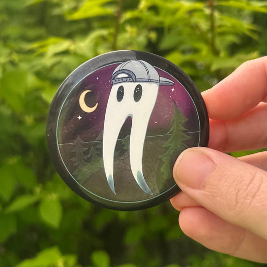 Hand holding a small round pin with a Fresno Nightcrawler design against a green outdoor background