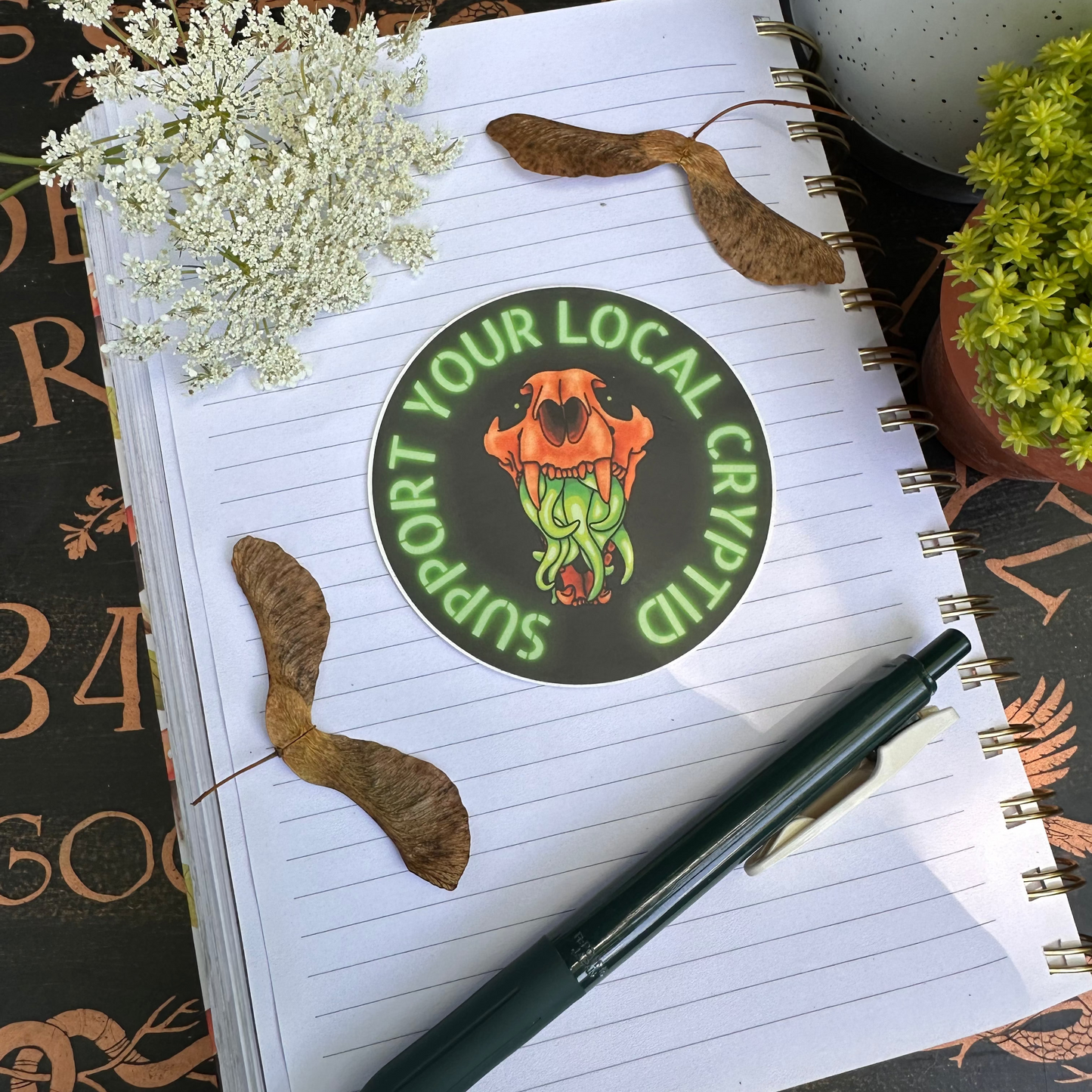 A sticker featuring a stylized illustration of an animal skull with vibrant green tentacles, and the text 'support your local Cryptid' around it.