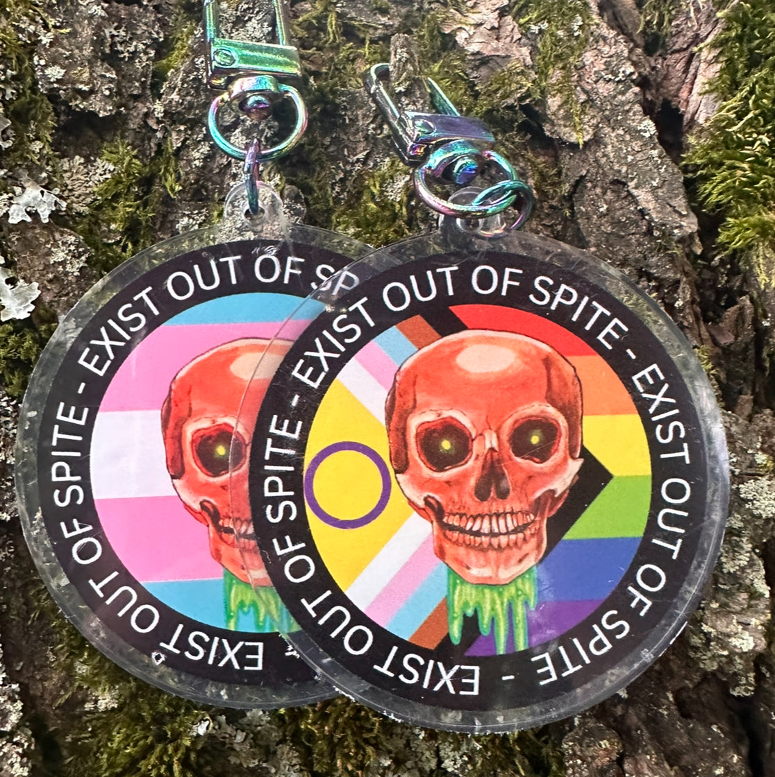 Two round keychains with a colorful skull design and text 'EXIST OUT OF SPITE' with a skull and pride flag on a textured surface.