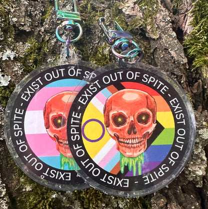 Two round keychains with a colorful skull design and text 'EXIST OUT OF SPITE' with a skull and pride flag on a textured surface.