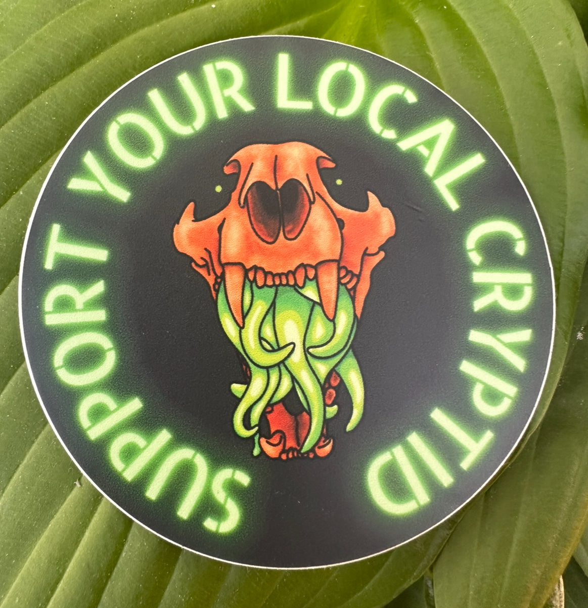 A sticker featuring a stylized illustration of an animal skull with vibrant green tentacles, and the text 'support your local Cryptid' around it.