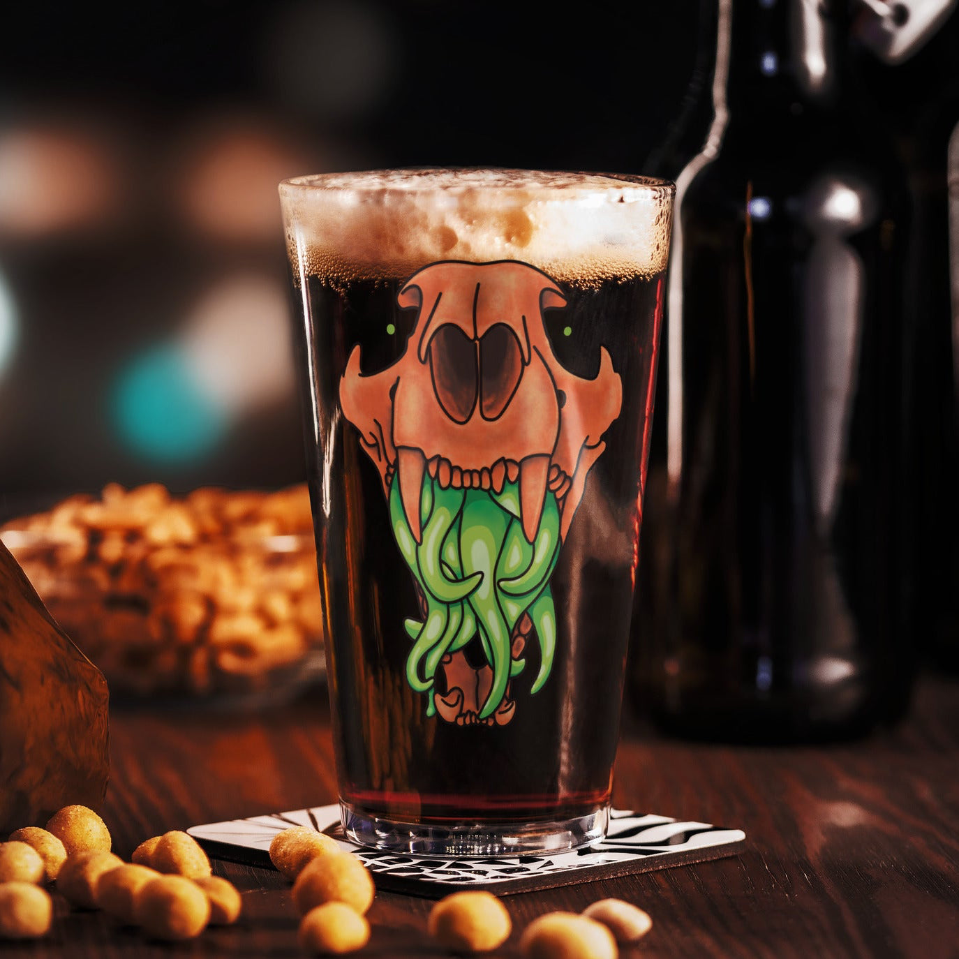 Pint glass with a decorative design of an animal skull with tentacles on a wooden table with beer bottles and snacks in the background.