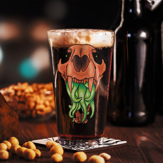 Pint glass with a decorative design of an animal skull with tentacles on a wooden table with beer bottles and snacks in the background.