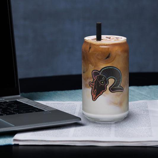 A glass mug with a design of a ram skull, filled with a beverage, placed on a table next to a laptop and a newspaper.