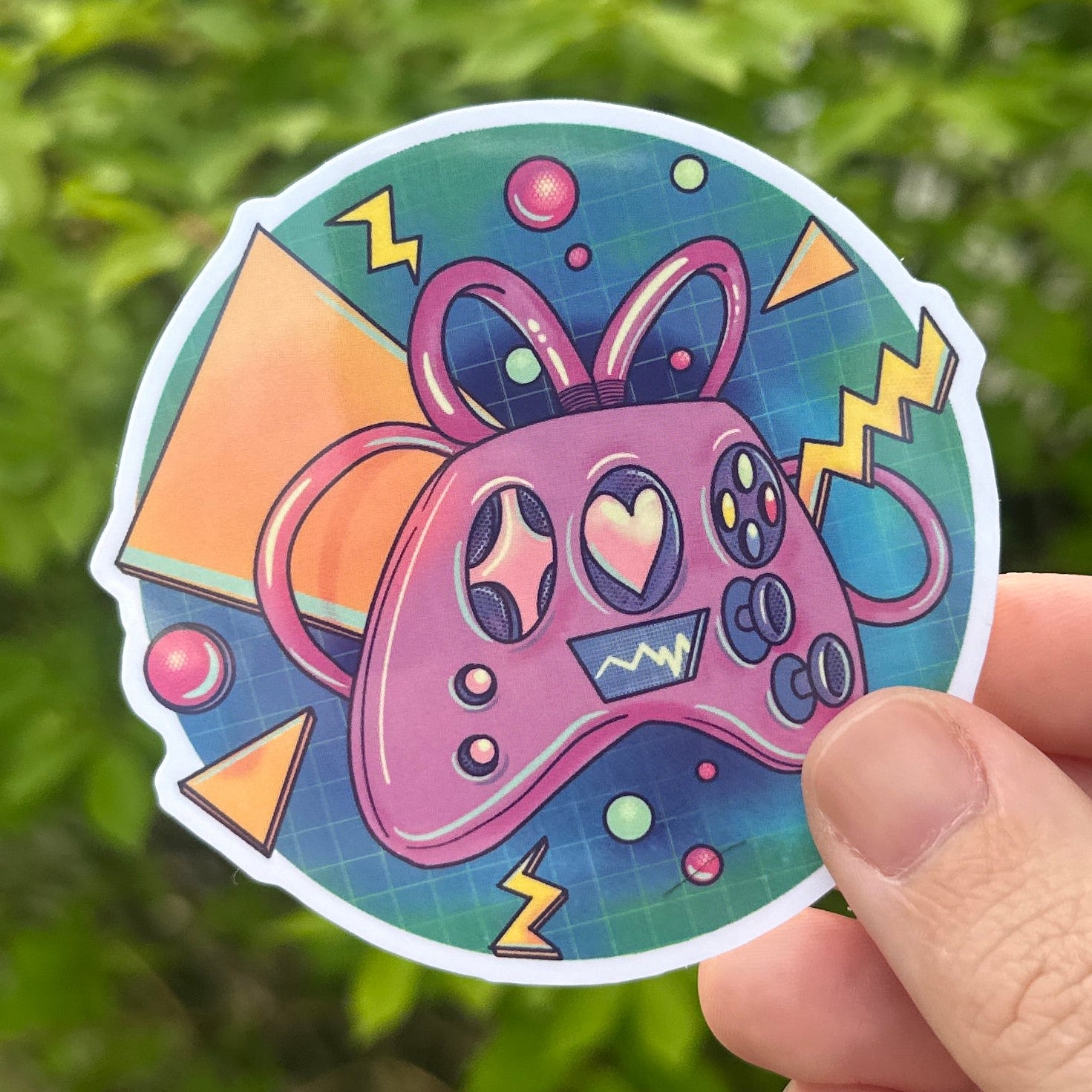 Colorful sticker with a retro controller design held by a hand against a green leafy background