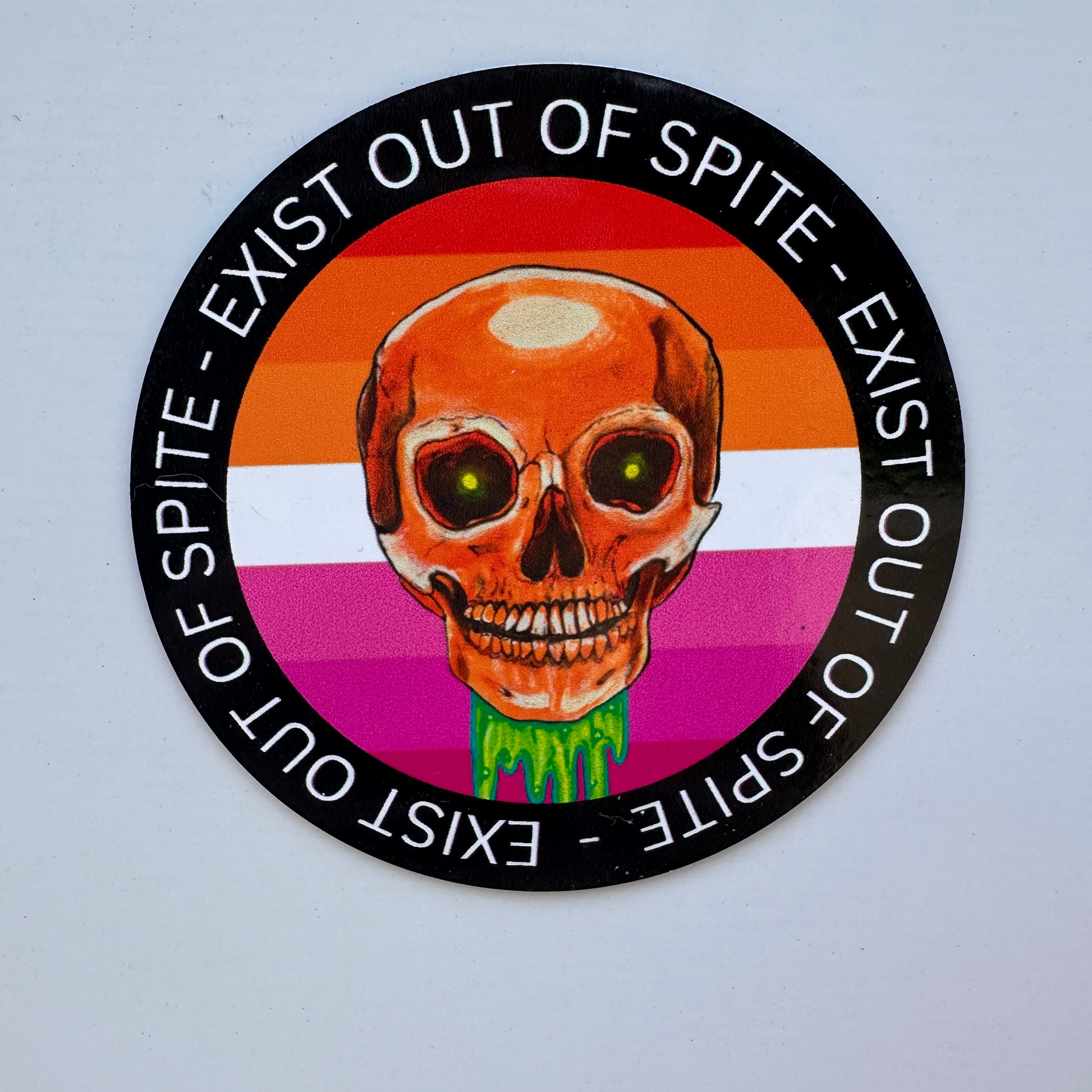 Round magnet with a skull over a lesbian pride flag encircled by the text exist out of spite