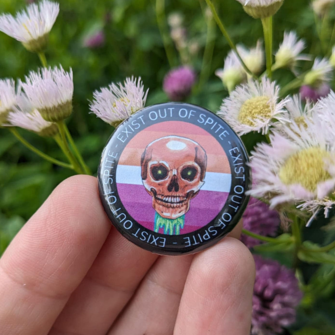 Round pin with a skull over a lesbian pride flag encircled by the text exist out of spite