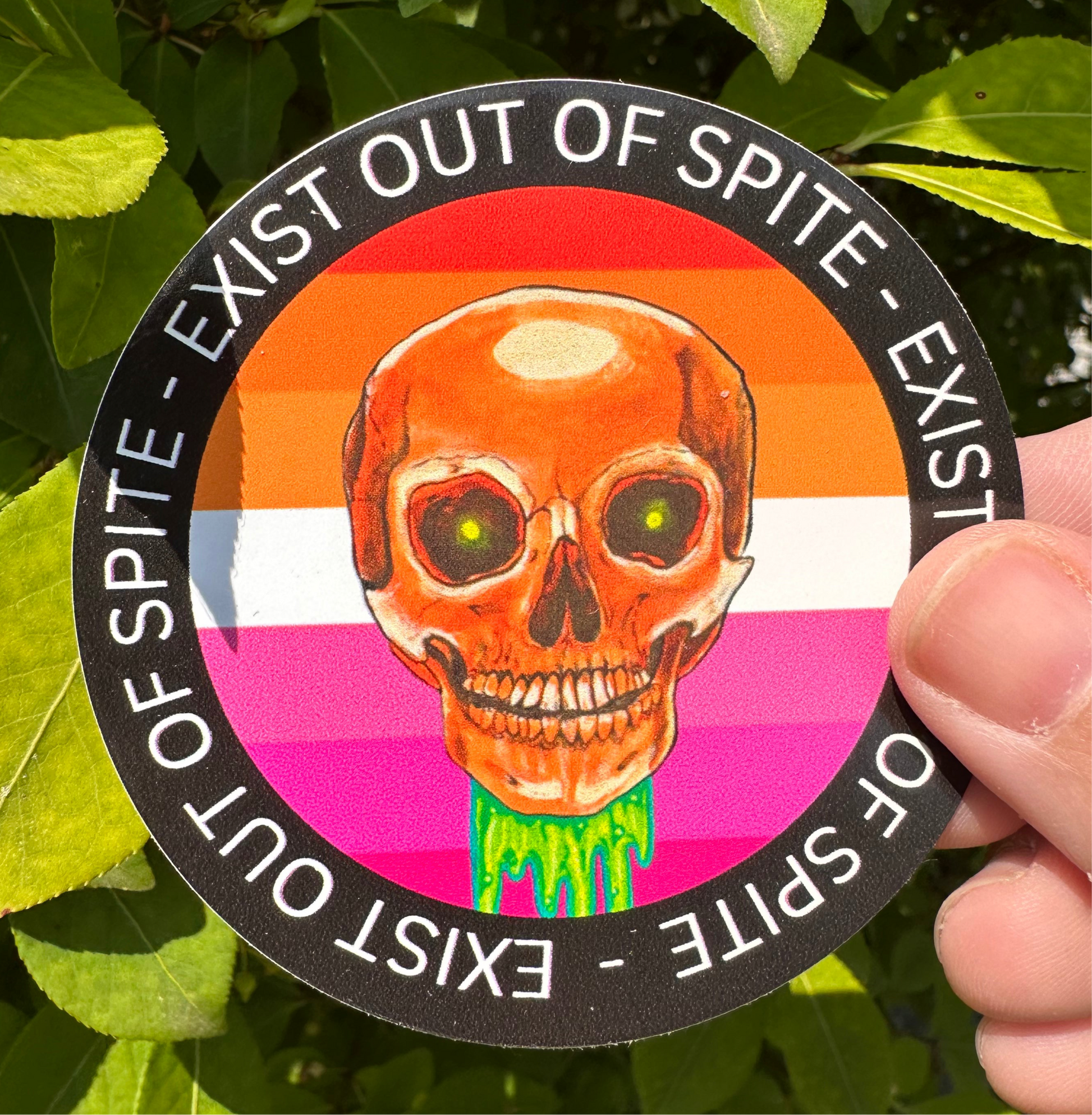 Round sticker with a skull over a lesbian pride flag encircled by the text exist out of spite