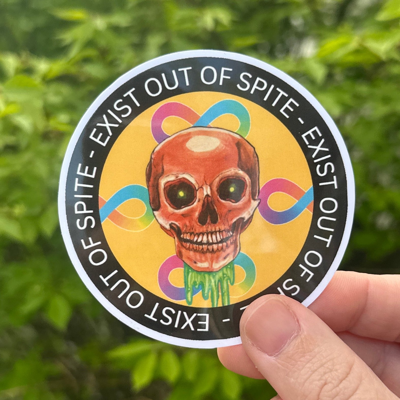 Hand holding a round sticker with a colorful design and text against a green leafy background