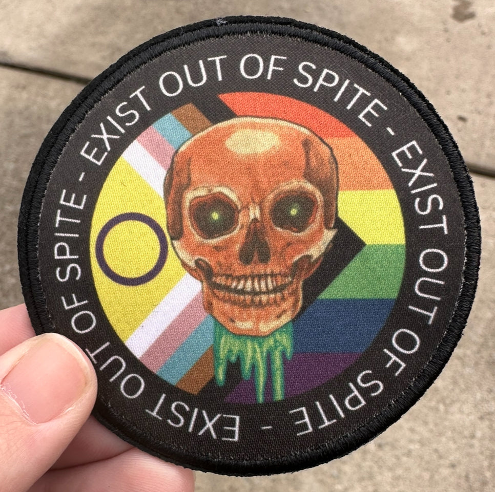 A circular patch featuring a skull design with the text 'EXIST OUT OF SPITE' arranged around it, set against a rainbow-colored background. The patch is held between fingers.