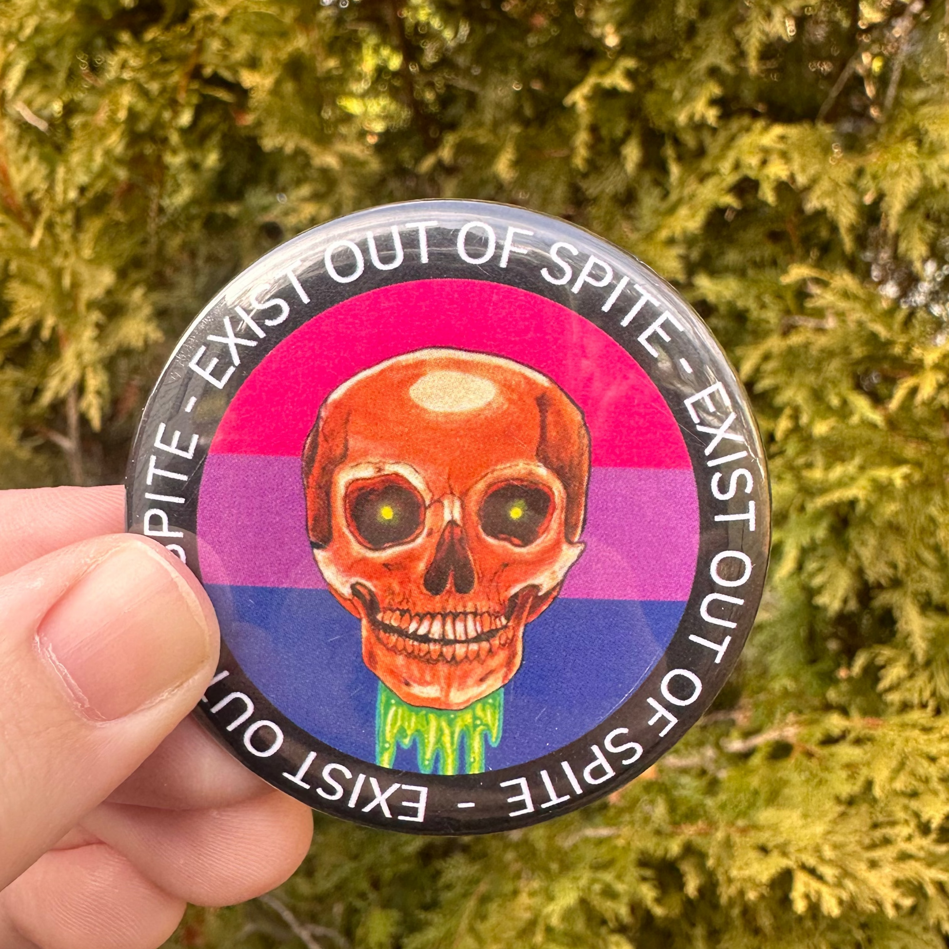 Round pin with a skull over a bisexual pride flag encircled by the text exist out of spite