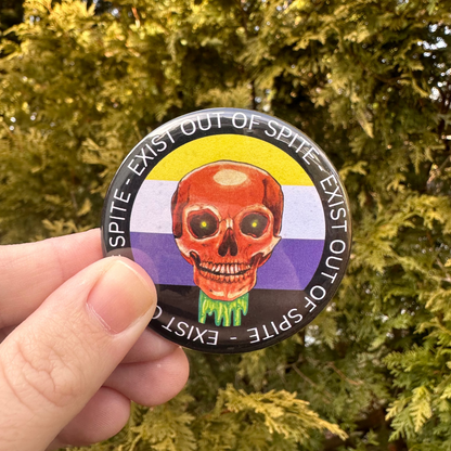 Round pin with a skull over a nonbinary pride flag encircled by the text exist out of spite