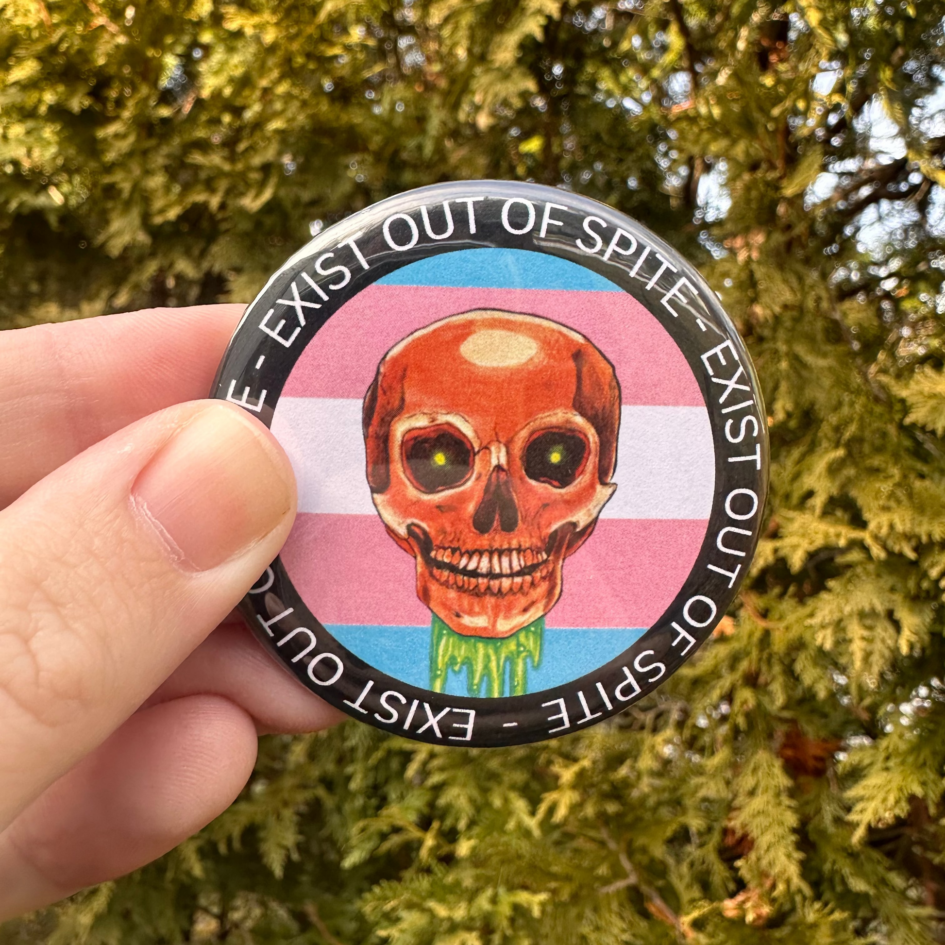 Round pin with a skull over a transgender pride flag encircled by the text exist out of spite