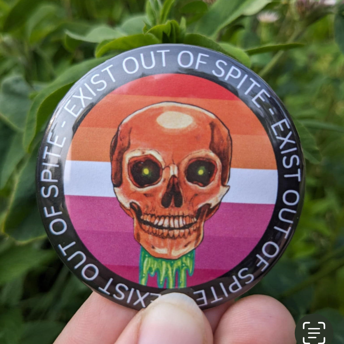Round pin with a skull over a lesbian pride flag encircled by the text exist out of spite