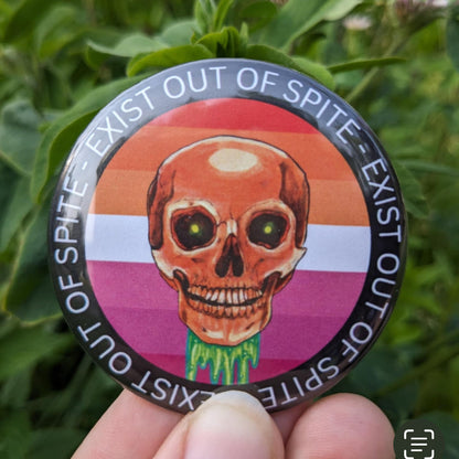 Round pin with a skull over a lesbian pride flag encircled by the text exist out of spite