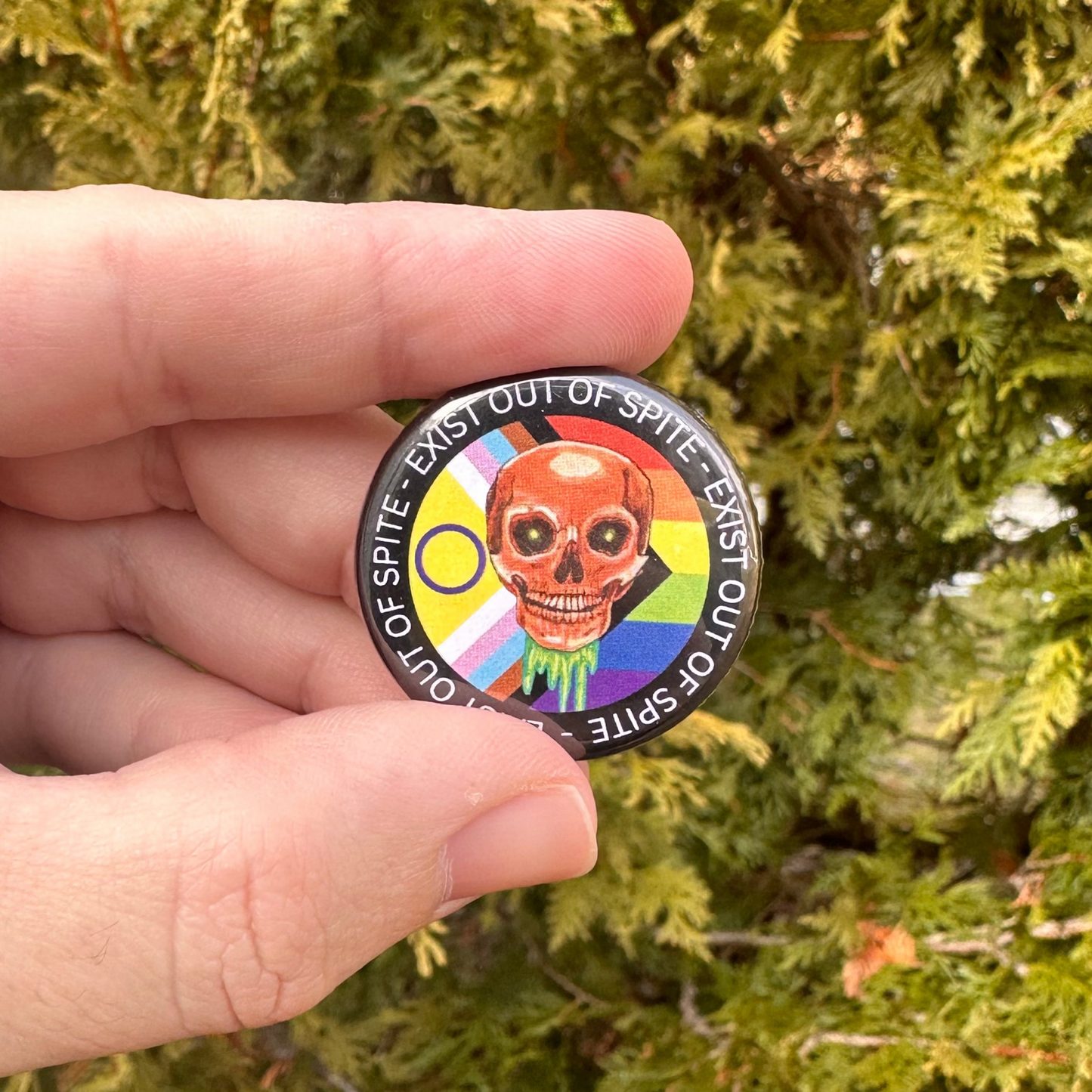 Round pin with a skull over a pride flag encircled by the text exist out of spite