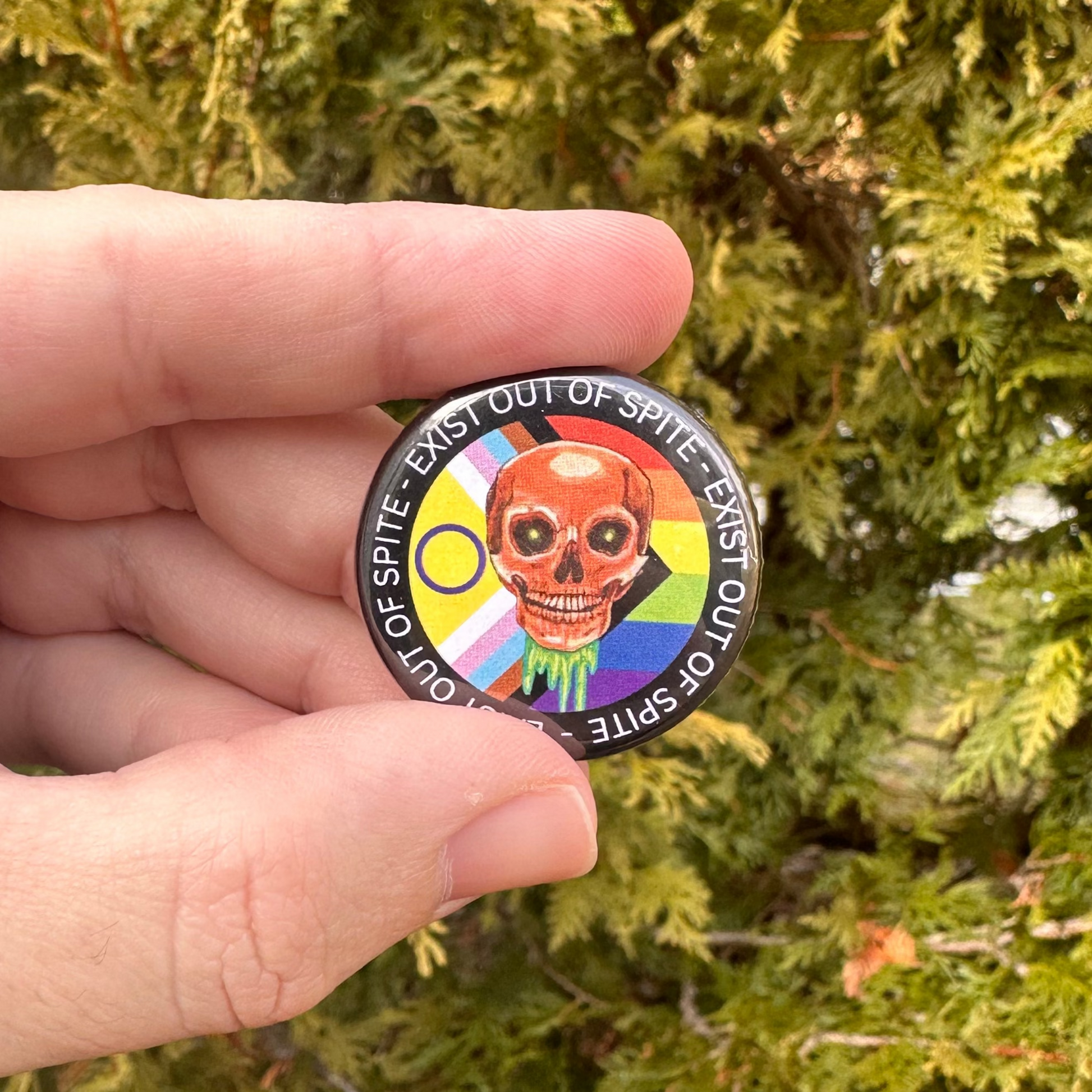 Round pin with a skull over a pride flag encircled by the text exist out of spite