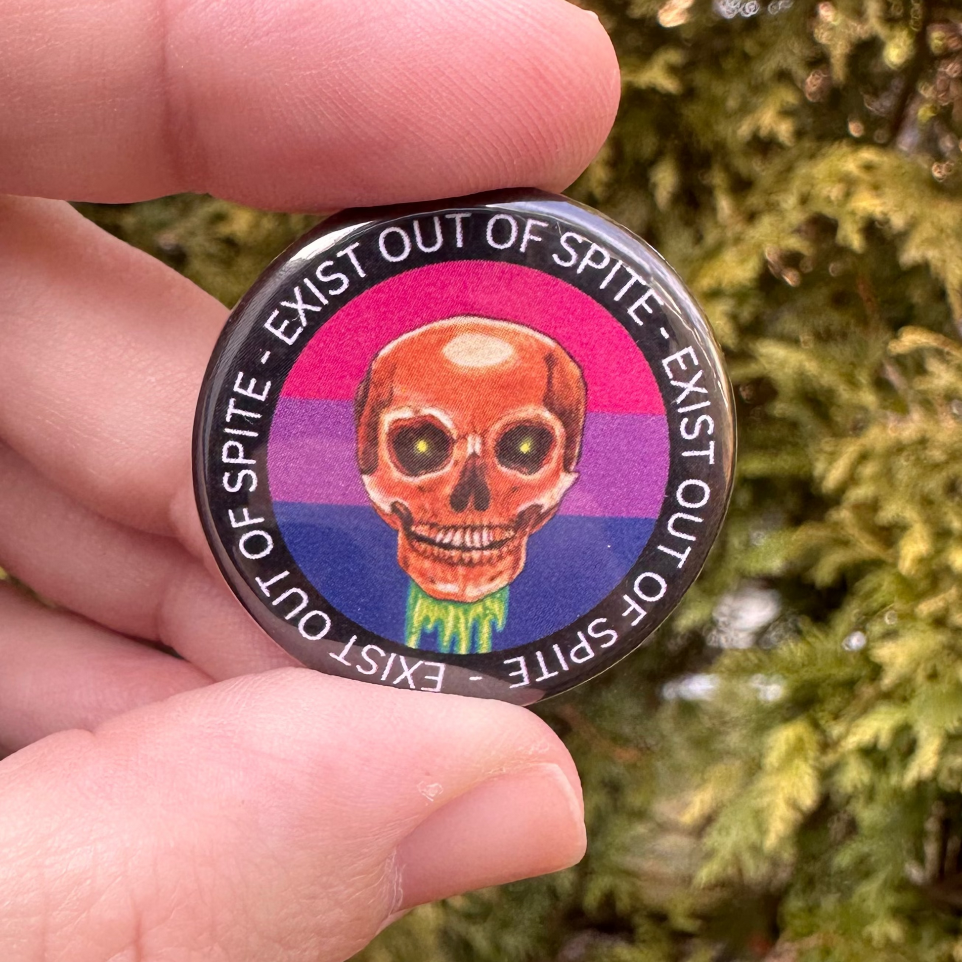 Round pin with a skull over a bisexual pride flag encircled by the text exist out of spite