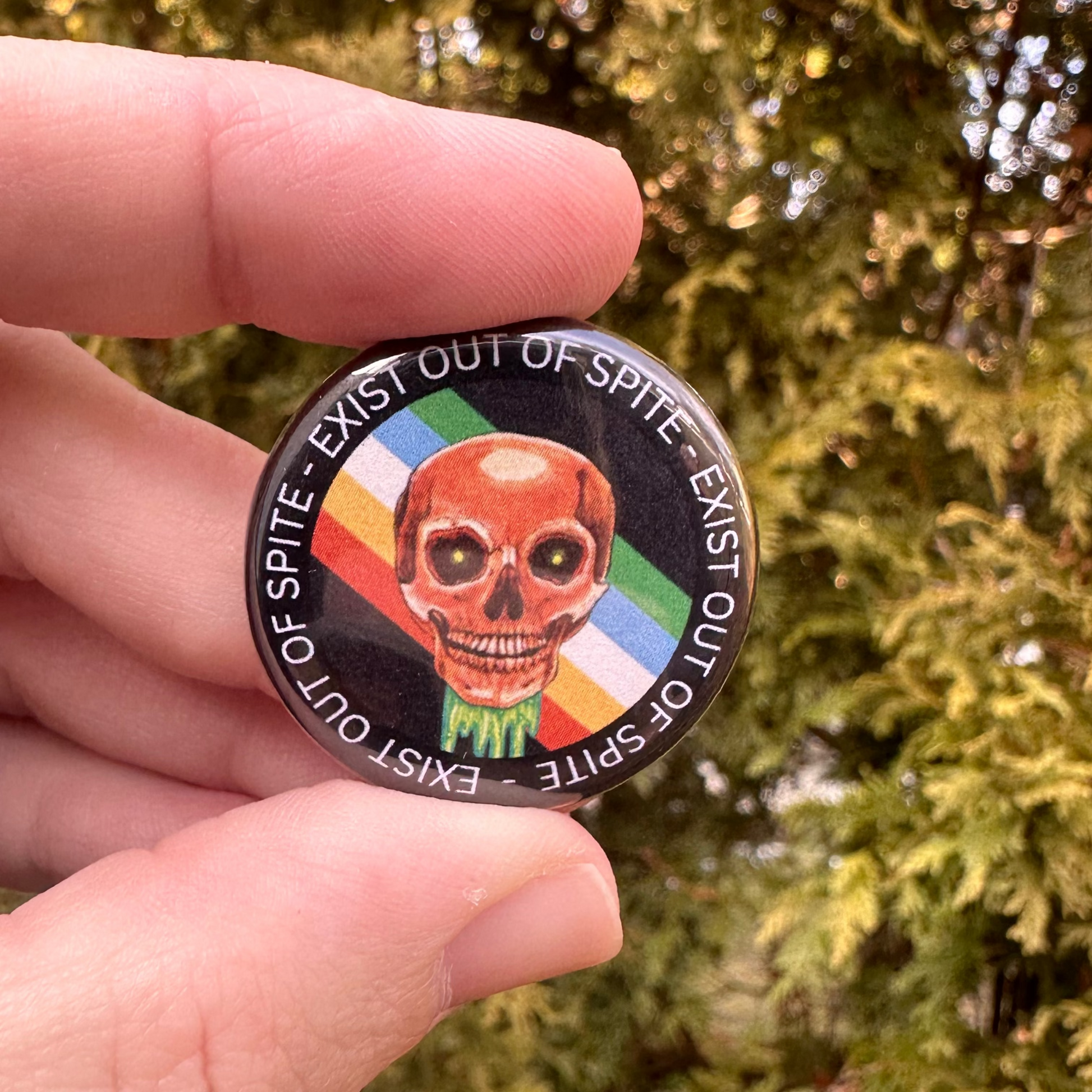 Round pin with a skull over a disabled pride flag encircled by the text exist out of spite