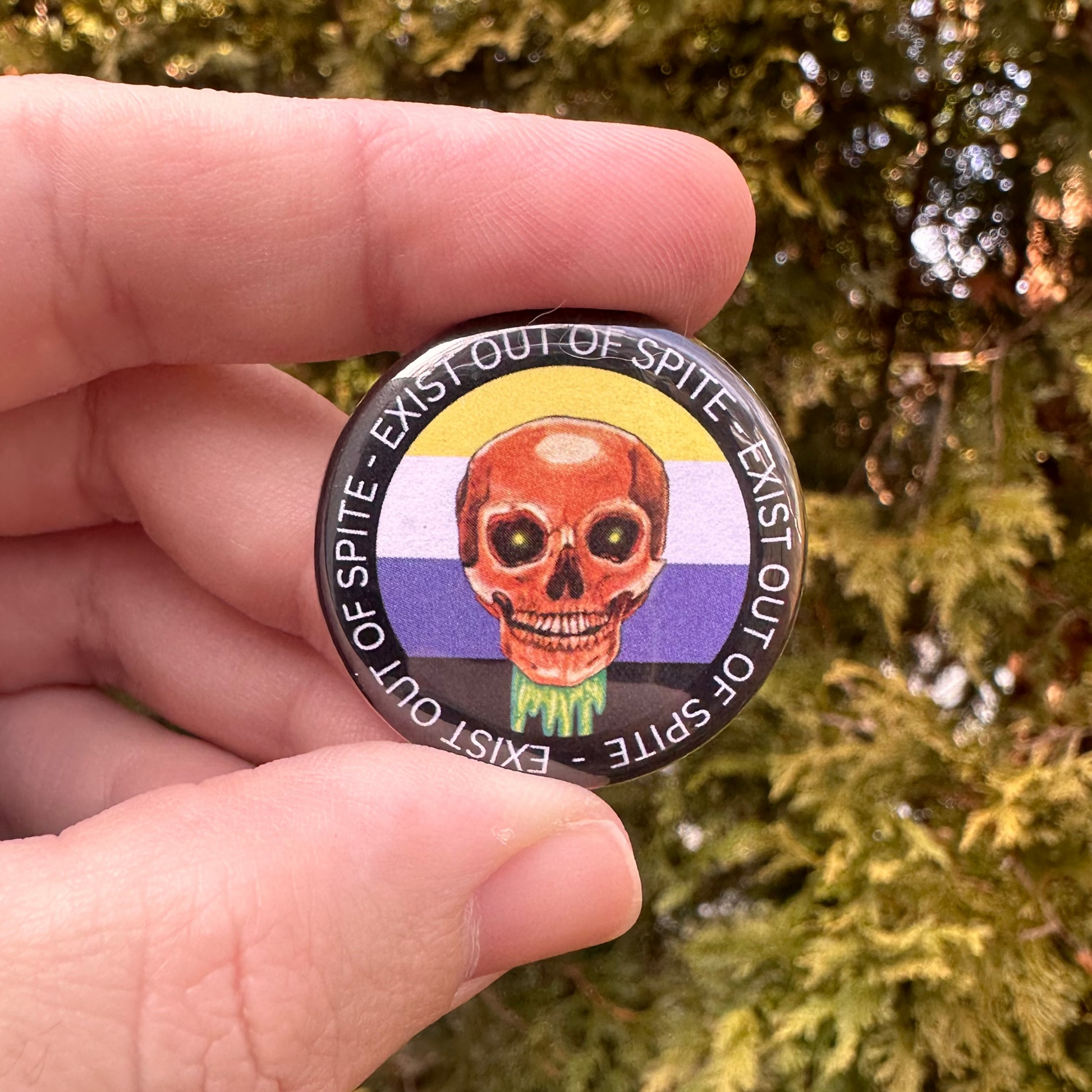 Round pin with a skull over a nonbinary pride flag encircled by the text exist out of spite