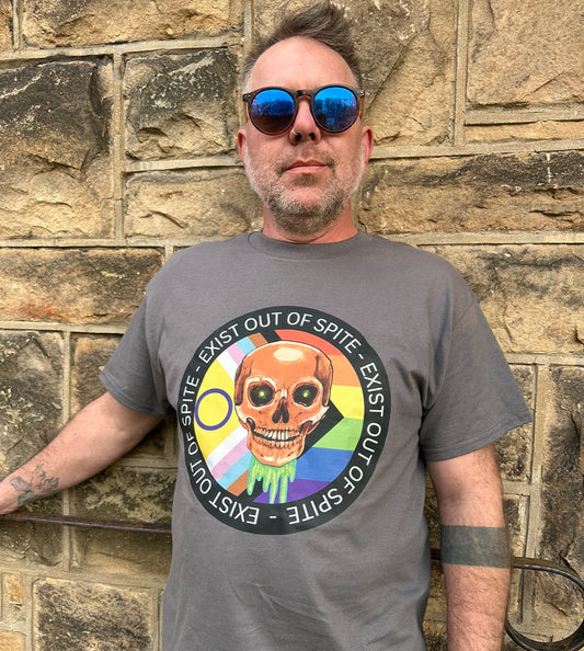 A person wearing a gray 'Exist Out of Spite' T-shirt with a colorful graphic of a skull and text over a pride flag on the front, standing against a stone wall background.