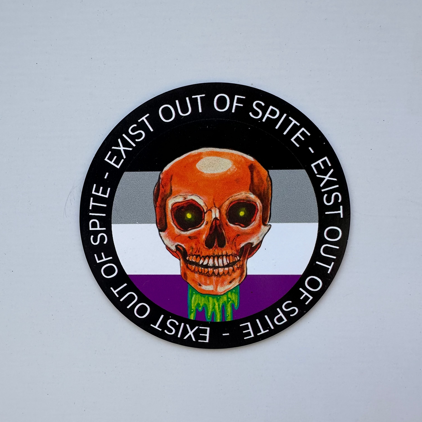 Round magnet with a skull over a asexual pride flag encircled by the text exist out of spite