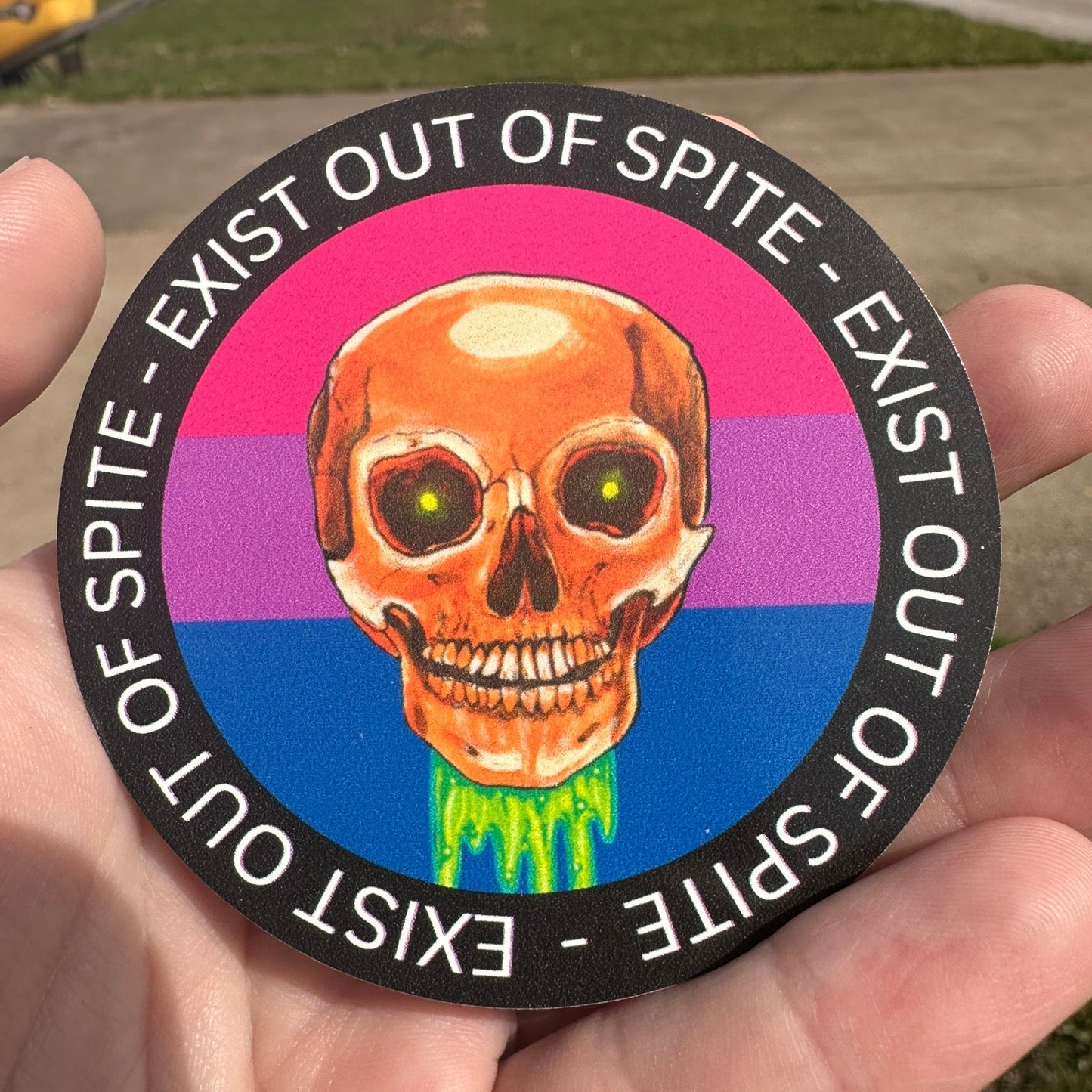 Round sticker with a skull over a bisexual pride flag encircled by the text exist out of spite