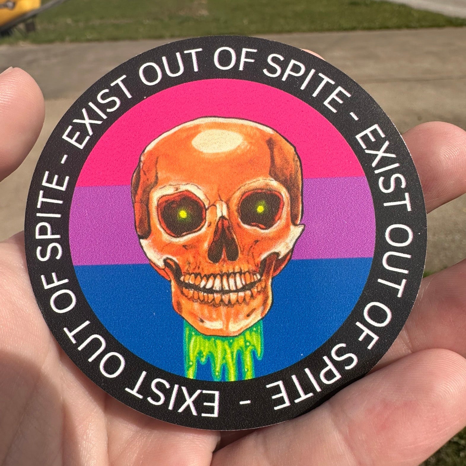 Round sticker with a skull over a bisexual pride flag encircled by the text exist out of spite