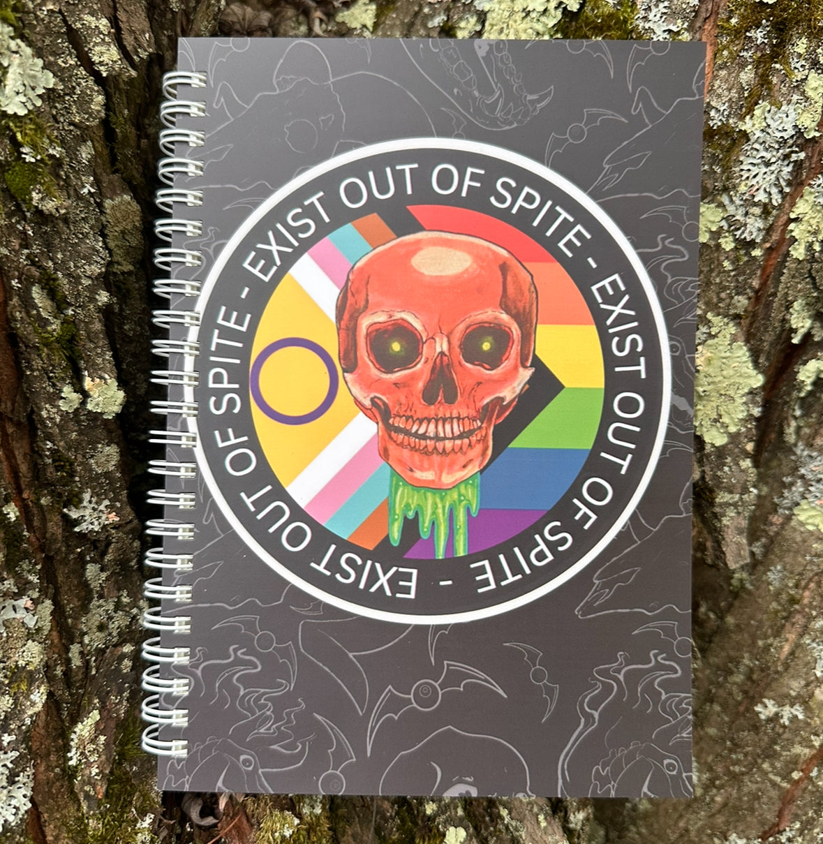 Journal with a circle design of a skull over a pride flag with text ‘exist out of spite’ circling on a subtle animal skull background