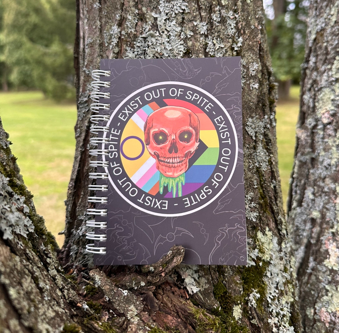 Journal with skull over a pride flag with text ‘exist out of spite’ circling on a subtle animal skull background