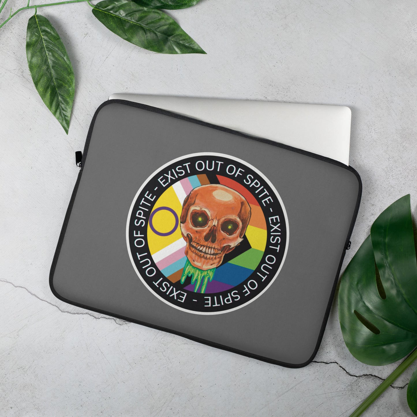 Laptop sleeve with a colorful skull design, pride flagand text ‘exist out of spite’ on a light gray surface with green leaves.