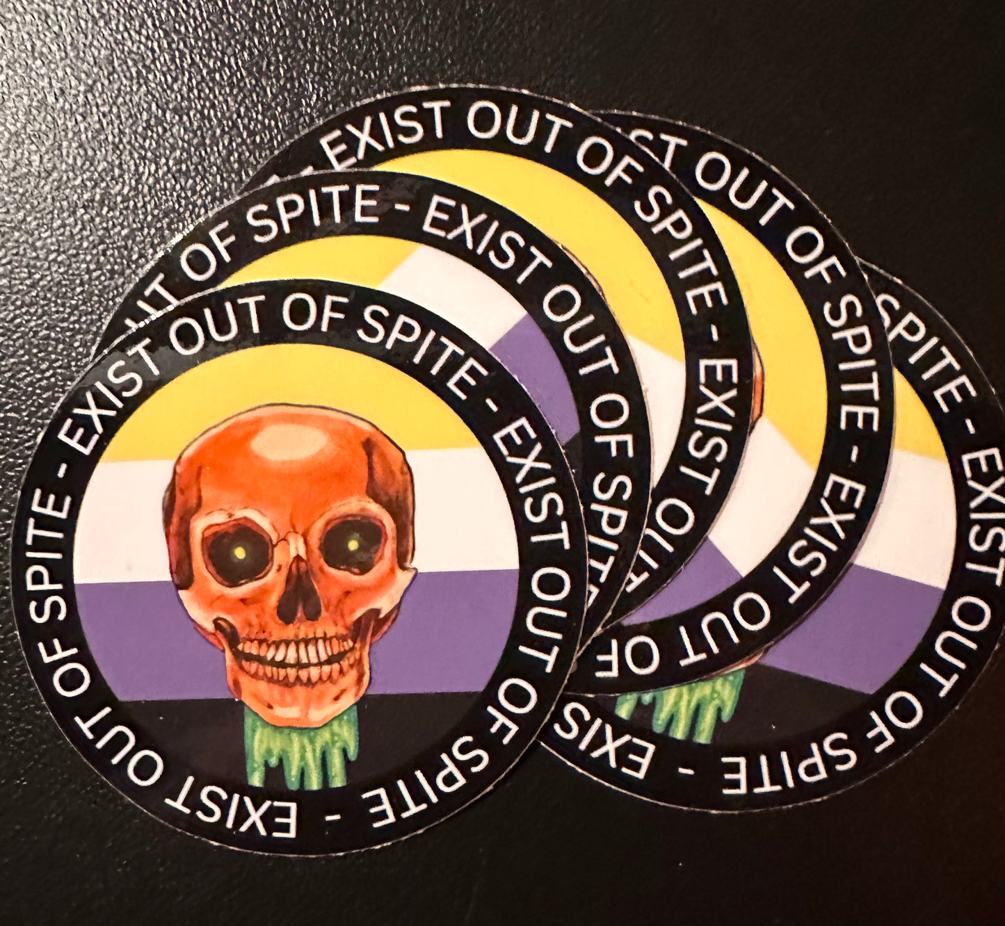 Round sticker 5 pack with a skull over a nonbinary pride flag encircled by the text exist out of spite